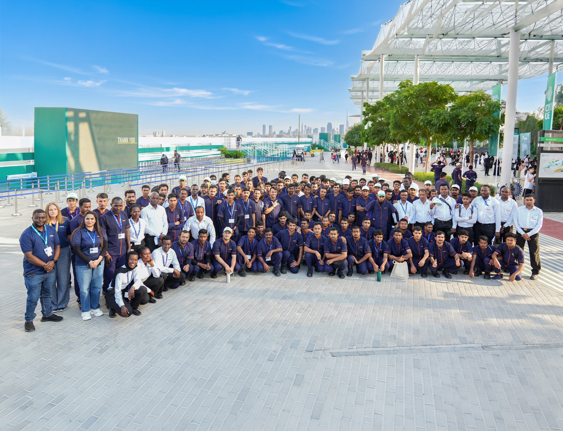 A large Farnek site team in matching navy and orange uniforms gathered on the plaza of a major Dubai event venue, with the city skyline behind